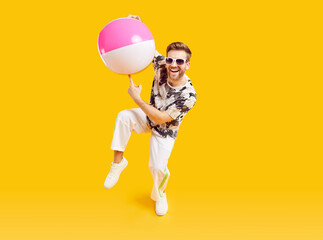 Funny young man with inflatable beach ball in sunglasses going on summer holiday trip. Happy guy tourist having fun isolated on studio yellow background. Vacation, journey and summertime concept.