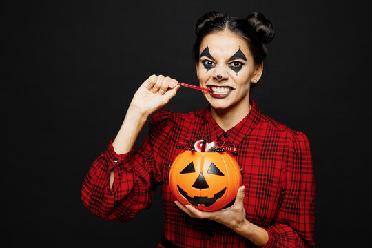 Young fun woman with Halloween makeup face art mask wear clown costume red dress hold Jack-o-Lantern carved pumpkin eat sweets isolated on plain black background studio. Scary holiday party concept.