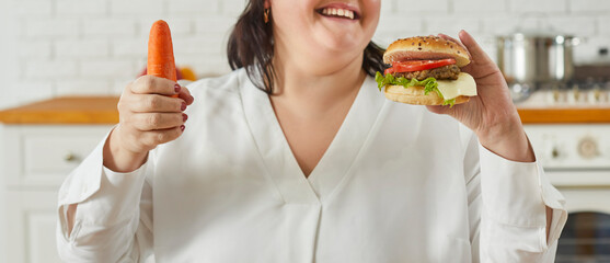 Overweight woman fails to start healthy diet. Fat woman with carrot and burger in hands. Happy lady chooses tempting tasty fast food hamburger. Crop shot banner. Healthy, unhealthy diet choice concept