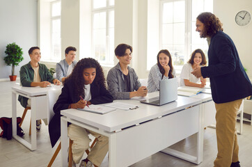 Male teacher conducts lesson for high school students and discusses topic of lesson with them. Smiling group of students sitting at desks in classroom and listening to teacher. Education concept.