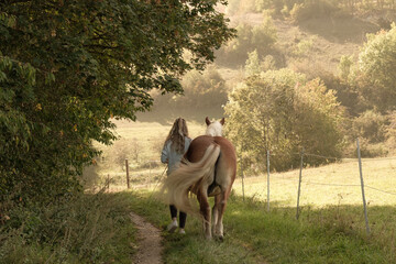 Frau / Mädchen mit Haflinger Pferd beim Spaziergang in der Natur auf einem Weg beim warmen Licht