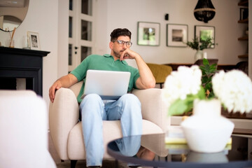 Thinking man sitting in an armchair at home and using laptop for work