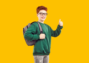 Fototapeta na wymiar Happy funny red-haired schoolboy with glasses, dressed in green sweater and gray jeans with backpack on his back shows thumbs-up - cool, photo on isolated yellow background. School education concept.