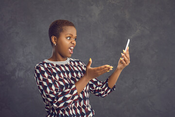 Portrait of dissatisfied and embarrassed dark-skinned woman who has problems with her mobile phone. Woman standing on gray background outraged by virus attack, lack of mobile communication or internet
