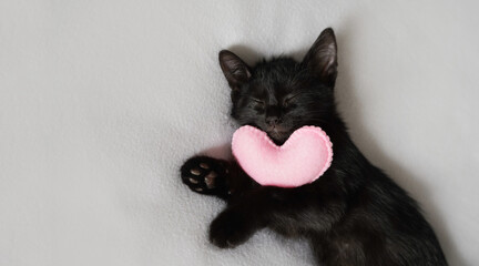 black kitten with a pink heart toy sleeping on a white background. portrait of a domestic kitten on white bed linen. Cozy house concept - Powered by Adobe
