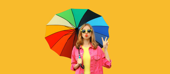 Autumn portrait of stylish young woman posing with colorful umbrella blowing her lips sends kiss wearing pink jacket on bright yellow studio background