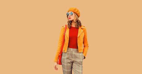 Autumn style outfit, beautiful stylish young woman posing with handbag looking away wearing orange french beret hat, jacket and round sunglasses on beige studio background