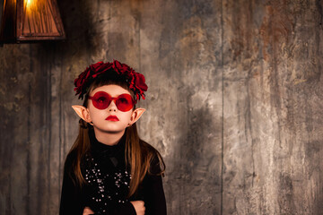 Portrait of mystic kid girl princess in blood glasses posing arms crossed at textured wall in dark room, bored looking up. Child actor in theatrical image. Theatre perform concept. Copy ad text space - obrazy, fototapety, plakaty