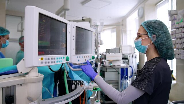 Female doctor wearing glasses is focused on the screens of lung ventilating machine. Anesthesiologist presses a button on the monitor. Surgeons work at backdrop.