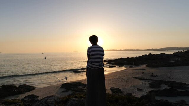 Video of a young boy sat on a bollard on the beach during the sunset, rear view, no faces are shown