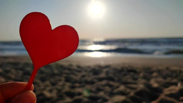 Woman holding in hand red stick in shape of heart on background of shining morning sun on sandy beach of sea shore near sea waves on sunny summer morning. Love romantic amorous St Valentines Day