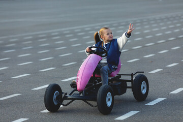 Little girl is riding on pedal karting. Child on a toy pedal car rides on a bike path outdoor,...