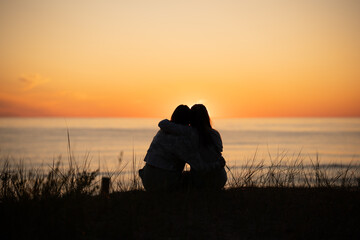 Silhouette of two female friends enjoying a conversation while sitting hugging and watching the sunset on a beach. Back view - obrazy, fototapety, plakaty