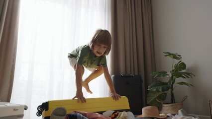 Child jumps on packed suitcase, rejoices, smiles. Small boy child throws plush toy into chaotically disorderly packed suitcase, climbs on top and jumps. Kid cheerful about vacation dancing happy dance - Powered by Adobe