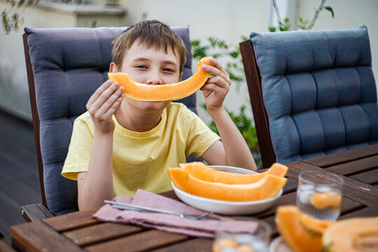Happy preteen boy sitting at table and eating fresh cut musk melon
