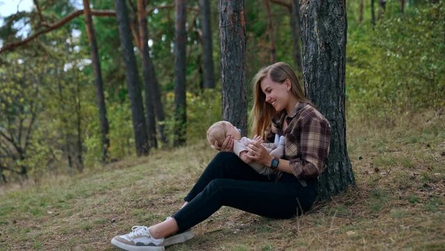 Happy long-haired Caucasian woman sits at the tree holding her baby on the laps. Mother talks and smiles to her adorable infant son.