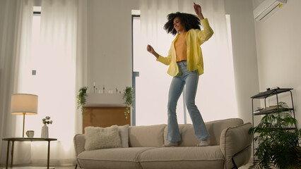 Funny African American woman happy girl at home dancing jumping have fun fooling joy testing comfortable soft couch cozy sofa furniture for apartment dance to music jump rejoice moving day relocation