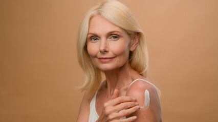 Mature old Caucasian woman in beige studio apply moisturizing body lotion sunscreen cream on...