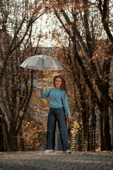 Young smiling woman with long hair poses in fall park. Woman in blue sweater and pants and holds transparent umbrella