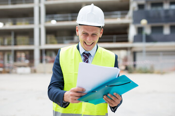 Positive young engineer standing on a construction site, attentive studies work documents