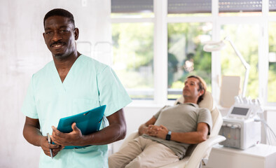 Fototapeta premium Middle-aged African man doctor stands with arms crossed on chest in hospital office, medical center. Modern medical care, tests, consultation