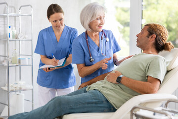 Obraz premium Senior woman physician talking to man lying on examination couch in medical office, discussing procedure details or treatment options. Female assistant filling out medical record