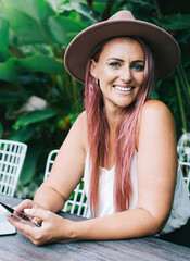 Fototapeta premium Cheerful gorgeous young woman sitting at table with laptop surfing mobile on cafe