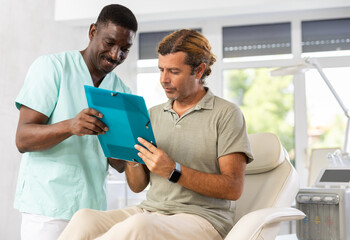 Experienced African American esthetician obtaining consent of male patient for hardware cosmetic procedures. Adult man reading and signing agreement while sitting on couch in aesthetic medicine clinic