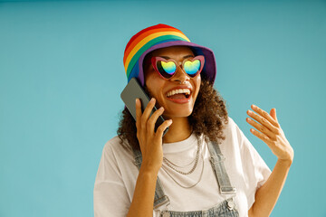 Cheerful woman in rainbow cap is talking by mobile phone standing over blue studio background