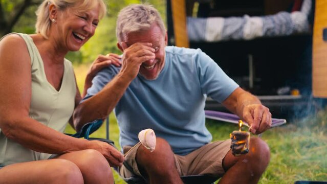 Senior retired couple camping in countryside in RV toasting marshmallows outdoors on fire with man burning his - shot in slow motion