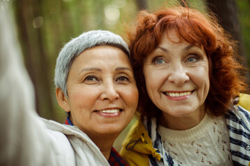 Obraz premium Multiracial elderly women having fun during trekking day in to the wood and make selfie with smartphone.