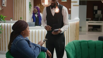 Woman does nfc mobile payment at pos to pay for cup of coffee while waiting on room reservation in hotel lounge area. African american tourist talking to porter about excellent concierge service.