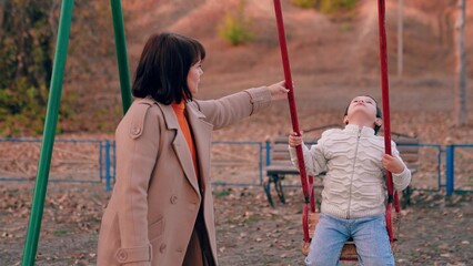 Happy family. Little son sitting on swing smiling, mom rocking child on children swing in park in fall. Autumn, little son plays on swing on playground under supervision of his mother. Mom kid play