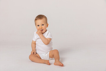 Little kid in white bodysuit, barefoot is smiling, sitting on floor against white studio background. Concept for articles about childhood or advertising for babies. Close up
