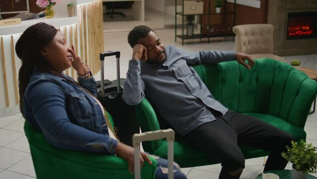 Tired partners having jet lag symptoms, napping with trolley bags in hotel reception entrance. Exhausted people travelling and feeling drained after long distance flight, waiting for check in process.