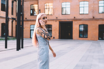 Stylish woman with tattoo speaking on smartphone in yard of administrative campus