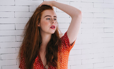 Confident woman with bright hair in red dress sitting near white brick wall