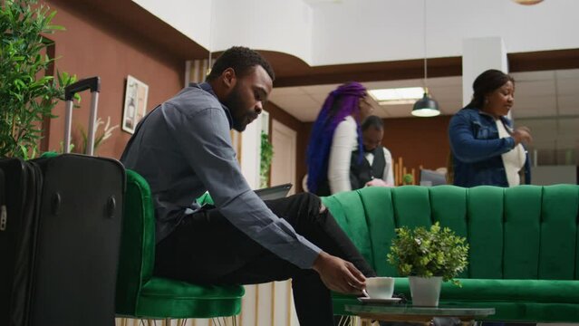 Hotel guest browsing tablet and enjoying coffee while he waits on room reservation check in, relaxing in lounge area at holiday resort. Young traveller receiving luxurious concierge services.