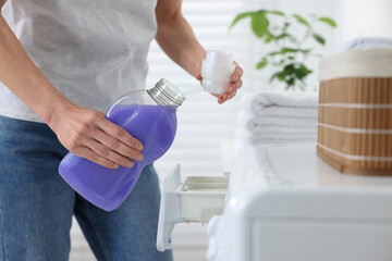 Woman pouring fabric softener from bottle into cap near washing machine in bathroom, closeup