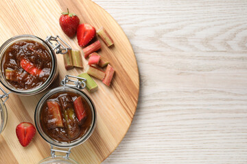 Jars of tasty rhubarb jam, cut stems and strawberries on white wooden table, top view. Space for text