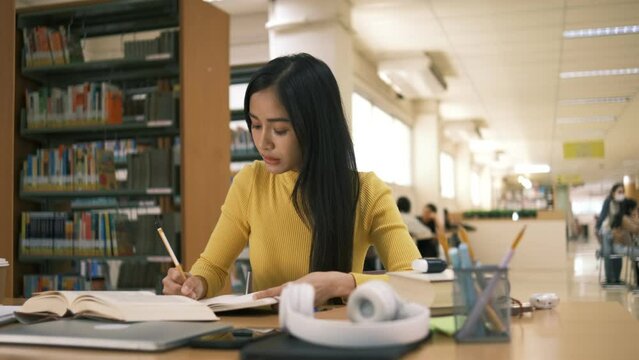 Asian student girl sitting at table looking book while do homework with laptop making video call abroad using internet friend connection, happy mood smiling broadly in library