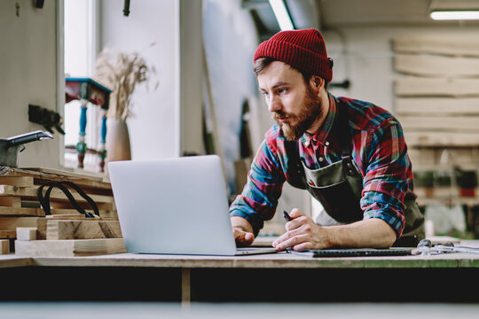 Creative male carpenter in apron browsing laptop at workshop