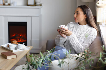 Young woman drinking cocoa with marshmallows near fireplace at home