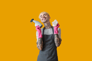 Young tattooed man with bottle of detergent and rag on orange background