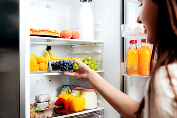 Woman hand taking, grabbing or picks up box of blueberry out of open refrigerator shelf or fridge drawer full of fruits, vegetables, banana, peaches, yogurt. Healthy food diet, lifestyle concept