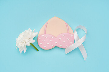 Pink cookie in shape of heart with bra, chrysanthemum flower and ribbon on blue background. Breast cancer awareness concept
