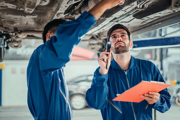 Car repair service. Two handsome young technician checking car condition in garage, Automotive mechanic pointing flashlight following at maintenance checklist document. Professional teamwork. - obrazy, fototapety, plakaty