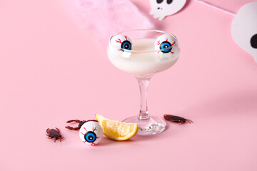 Glass of delicious white cocktail, lemon piece, eyes and candy bugs for Halloween celebration on pink background