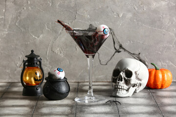 Glass of delicious cocktail, lantern, skull and eyes for Halloween celebration on grey background