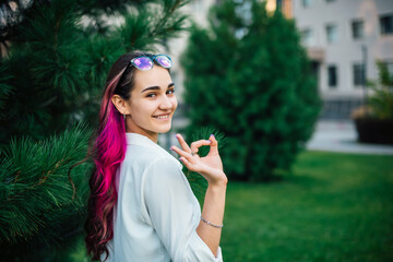 Portrait of smiling brunette with purple hair young woman makes okay hand gesture, indicates positive emotions. Smart female student in glasses gesturing everything is fine